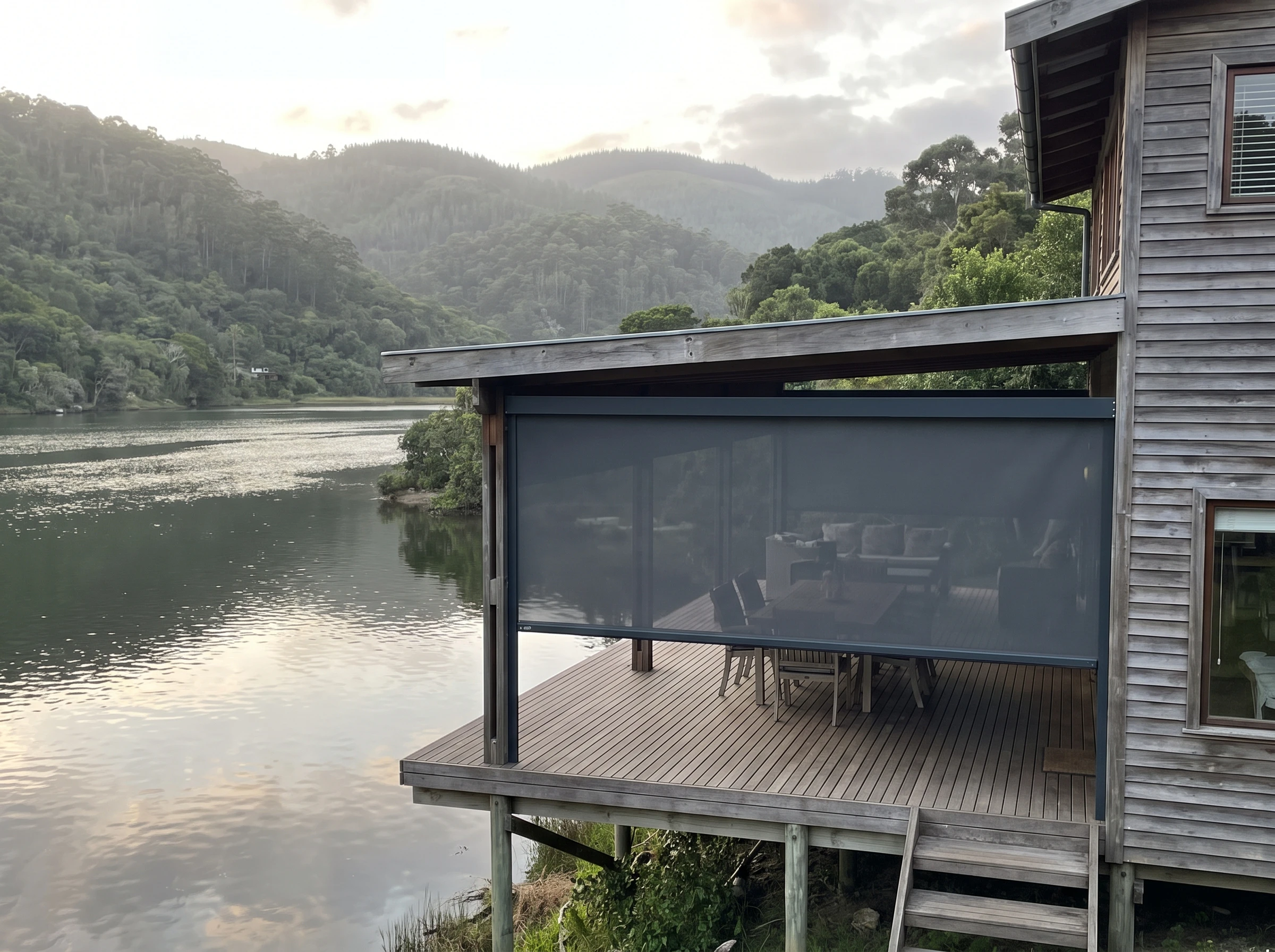 Timber-clad Garden Route home on the edge of the Knysna lagoon with forested hills behind and an outdoor zip screen partly closed on the covered deck